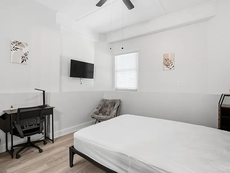 Private bedroom with bed frame, dedicated desk setup, wall-mounted TV, and a window with blinds for light control.