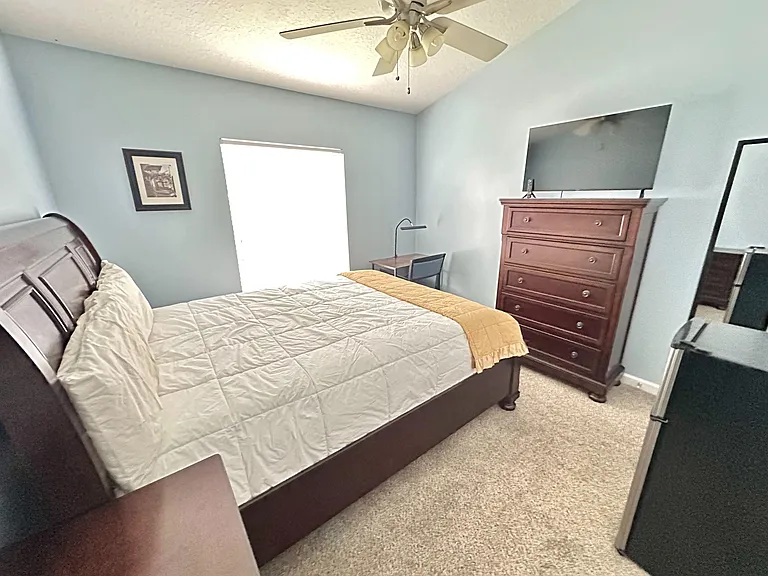 Furnished bedroom with a queen bed, dresser with TV, and a compact desk area, plus a ceiling fan and window for light.