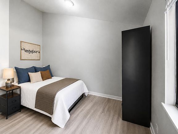 Private bedroom with a full-size bed, bedside table, and large wardrobe, plus a window and durable wood-look flooring.
