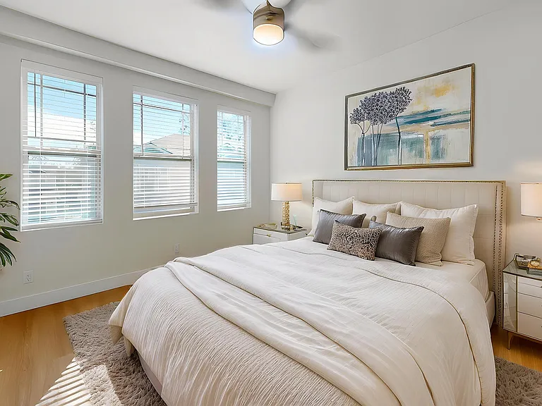 Sunlit bedroom with elegant decor, featuring a plush bed, stylish side tables, and modern artwork, complemented by natural light streaming through large windows.