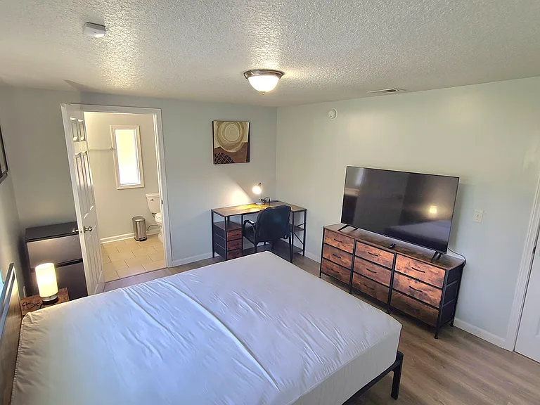 Furnished bedroom with a full bed, dresser with TV, and dedicated desk workspace, plus convenient access to a nearby bath.