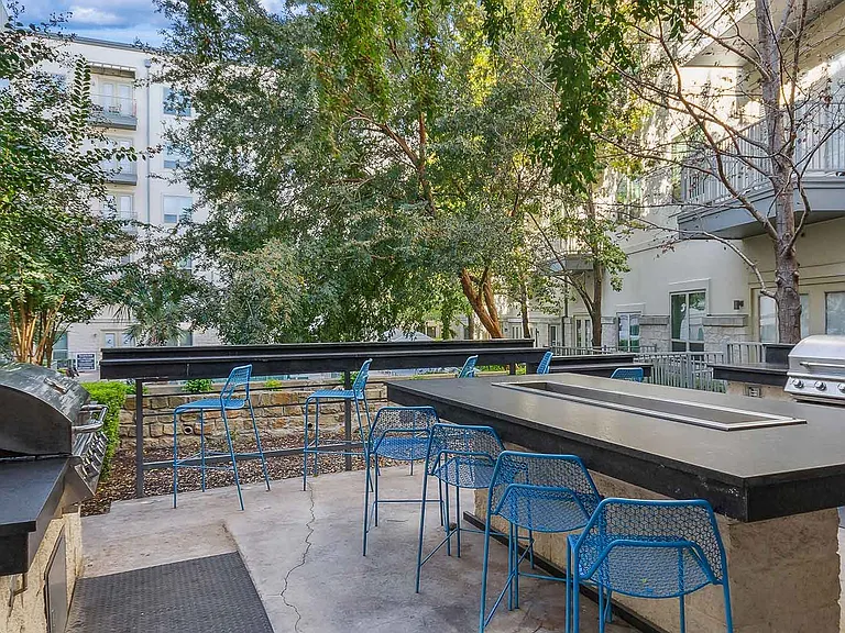 An outdoor grilling area at a residential complex, featuring modern stainless steel grills and a large stone countertop with a built-in dining surface. Vibrant blue bar stools surround the counter, of