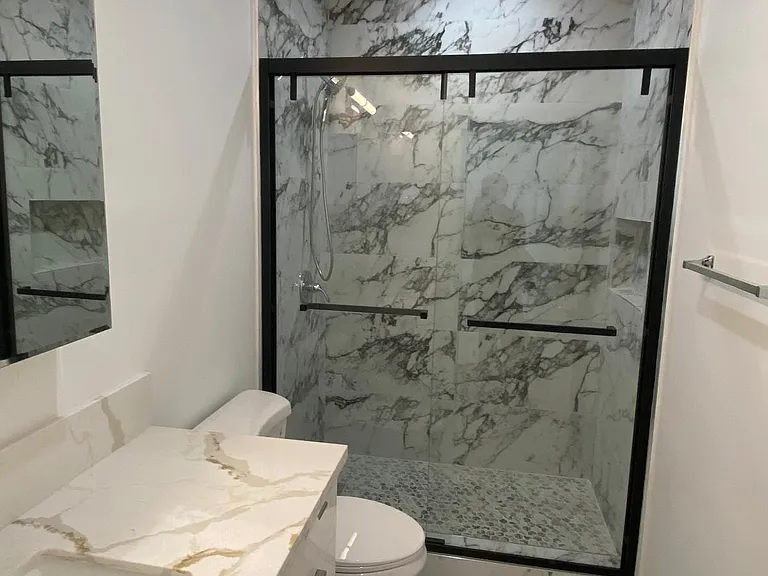 Modern bathroom with white vanity, marble-look countertop, large mirror, and glass-enclosed shower with marble-style tile.