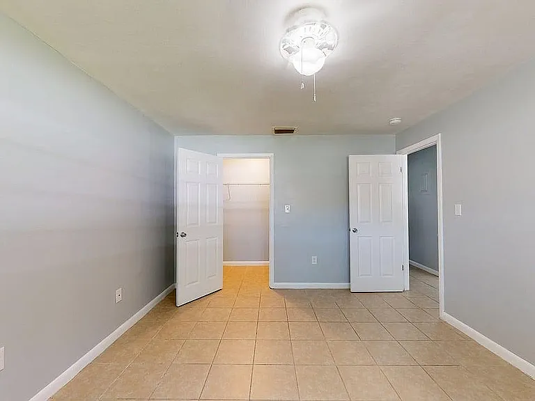Bright empty room with light gray walls, tiled flooring, and a ceiling fan, featuring two white doors leading to a walk-in closet and adjacent room.