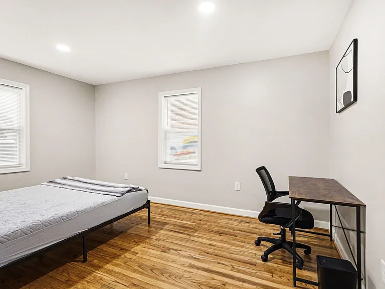 Furnished bedroom with hardwood floors, two windows with blinds, and a dedicated desk workspace for studying or WFH.