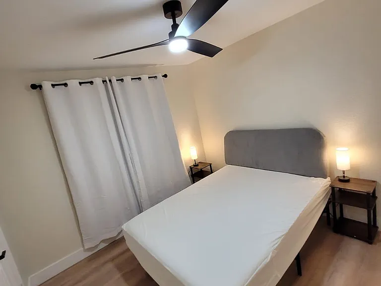 Furnished bedroom featuring a queen bed, nightstands with lamps, ceiling fan, and window with curtains over wood-look floors.