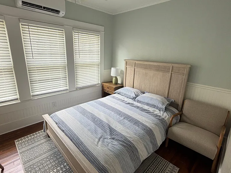 Furnished bedroom with a queen bed, nightstand, and seating area, plus ceiling fan, AC, and large windows with blinds.