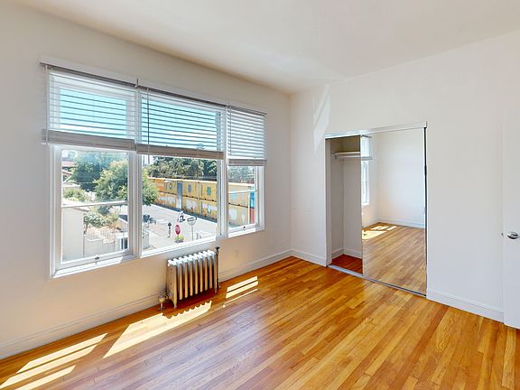 Bright, spacious bedroom with hardwood floors, large windows, and a mirrored closet reflecting natural light, street view visible outside