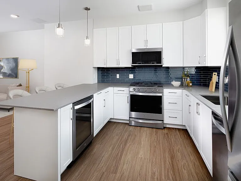 Phase II Renovated Package I townhome kitchen with stainless steel appliances, grey quartz countertops, white cabinetry, grey tile backsplash, and hard surface flooring