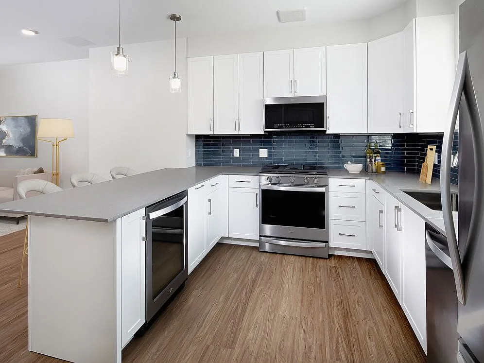 Phase II Renovated Package I townhome kitchen with stainless steel appliances, grey quartz countertops, white cabinetry, grey tile backsplash, and hard surface flooring - Avalon Acton