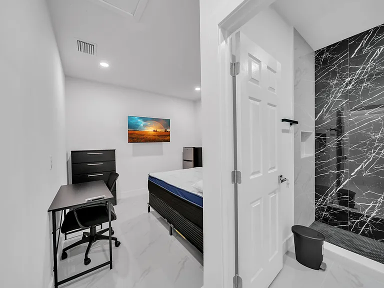 Private furnished bedroom with a work desk, dresser storage, mini fridge, and access to a sleek tiled ensuite shower.