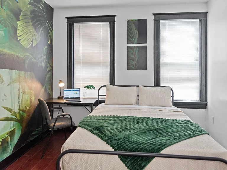 Furnished bedroom featuring a queen-size bed, dedicated work desk, and two large windows with blinds for privacy.