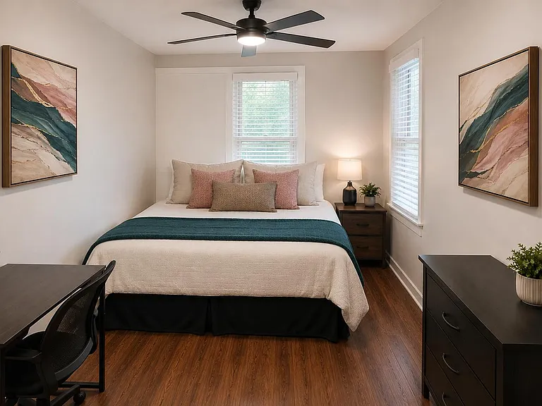 Spacious bedroom with a queen bed, hardwood floors, ceiling fan, dual windows, and dedicated desk plus dresser storage.