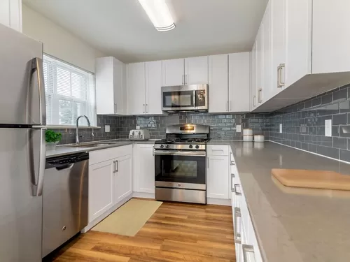 Renovated Package II kitchen with white cabinetry, grey quartz countertops, grey tile backsplash, stainless steel appliances, and hard surface plank flooring throughout common areas. Soft-close cabinets and microwaves available in select homes - eaves Wilmington and Wilmington West