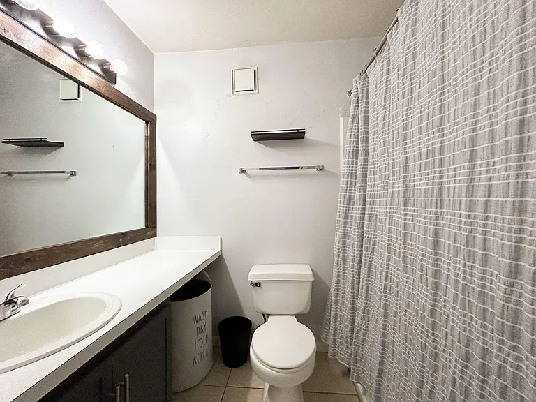 Full bathroom with long vanity counter, framed mirror lighting, and a tub/shower combo with curtain plus wall shelving.