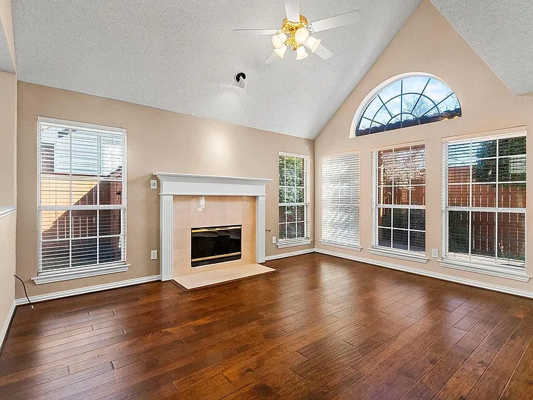 Vaulted ceiling with an arched transom window, wood-finish flooring, a fireplace with a light-toned tile surround and white mantel, and multiple windows with
 blinds