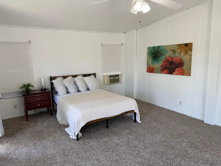 Furnished bedroom with a comfortable bed, carpeted floors, wall art, and two windows plus a window A/C unit.