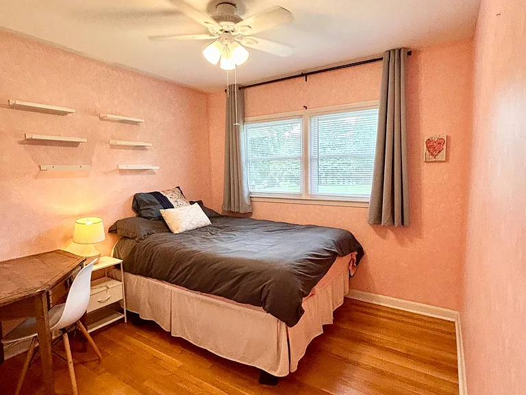 Furnished bedroom with hardwood floors, ceiling fan, and a large window with curtains plus a desk and wall shelving.