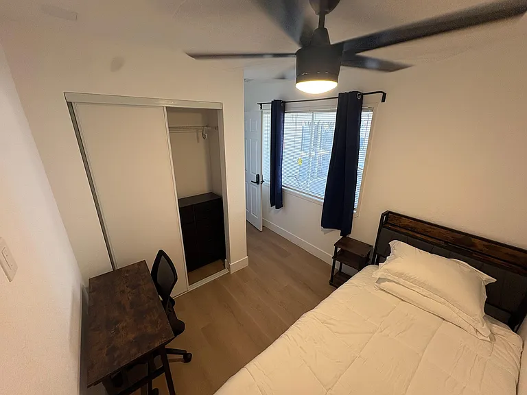 Furnished bedroom with a ceiling fan, window with blinds and curtains, desk area, and a sliding-door closet for storage.