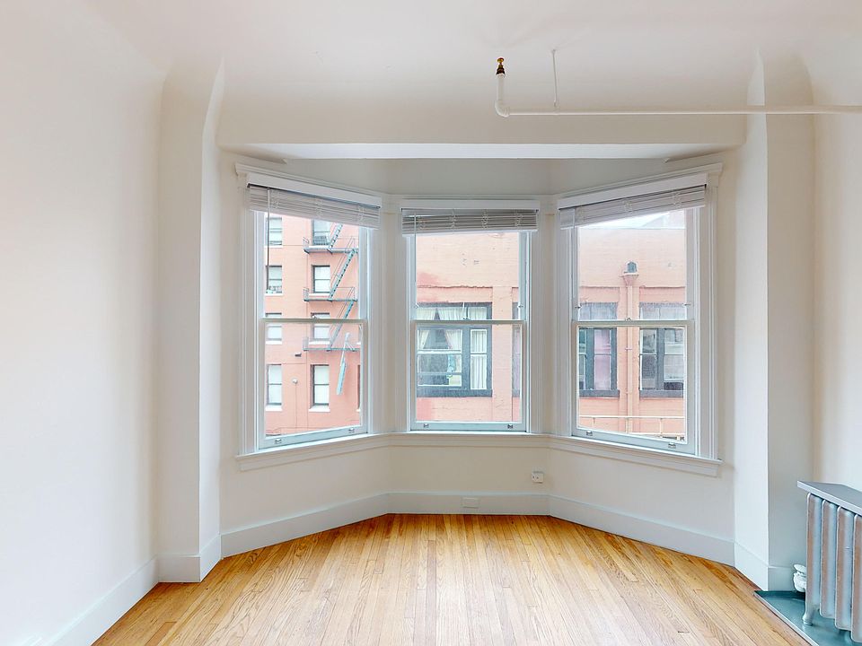 Bright, empty room with hardwood floors and a bay window offering a view of brick buildings. Simple, minimalistic, and spacious atmosphere.