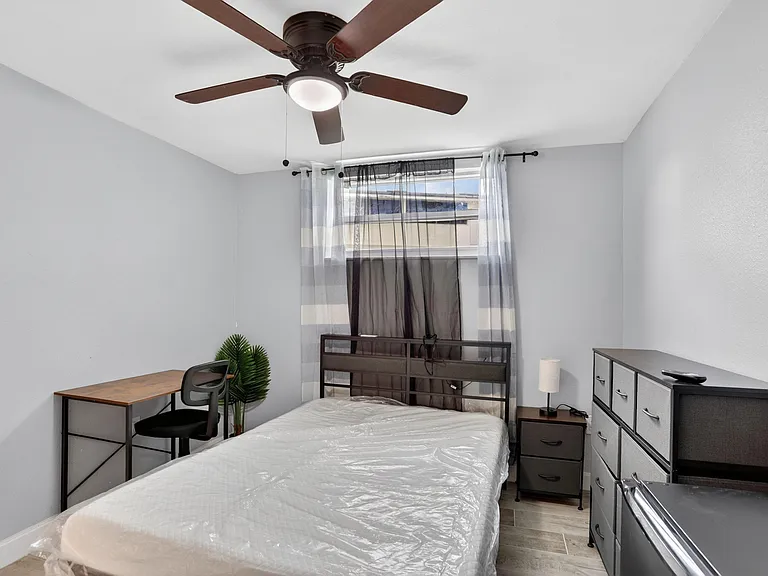 Furnished bedroom with a bed, dresser, nightstand, and dedicated desk area, plus a ceiling fan and window coverings.
