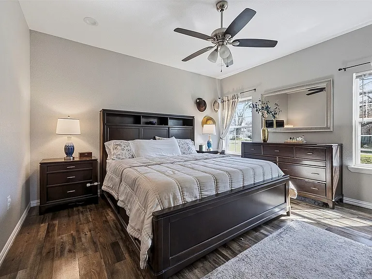Spacious bedroom featuring wood-finish flooring, two large windows, a ceiling fan with integrated lighting, and neutral wall paint