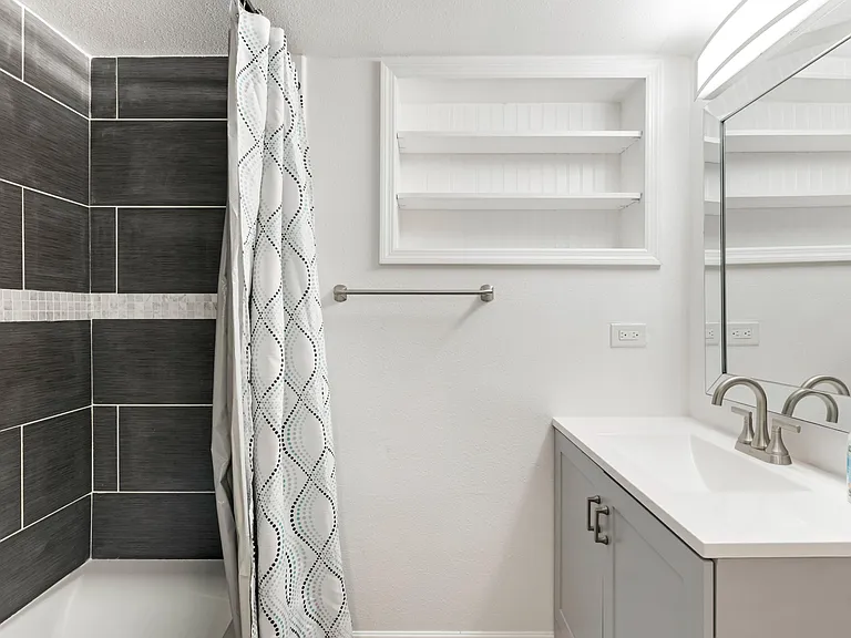 Updated bathroom with tiled tub/shower, single-sink vanity, mirrored cabinet, and built-in wall shelving for essentials.