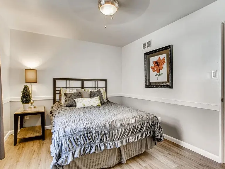 A bedroom with a bed, a small table with a plant, and a framed picture of a flower.