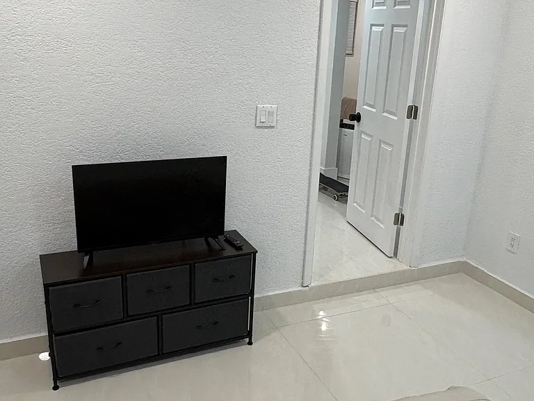 Bedroom features tile flooring, a dresser with TV, and easy access to the adjacent room through a hallway entry.