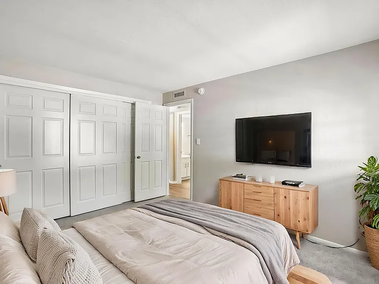 Inviting bedroom with plush bedding, modern TV setup, and stylish decor featuring a wooden dresser and lush green plant.