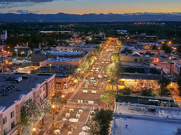 Enchanting evening aerial view of a lively city street lined with twinkling lights, bustling cafes, and picturesque mountain backdrop.