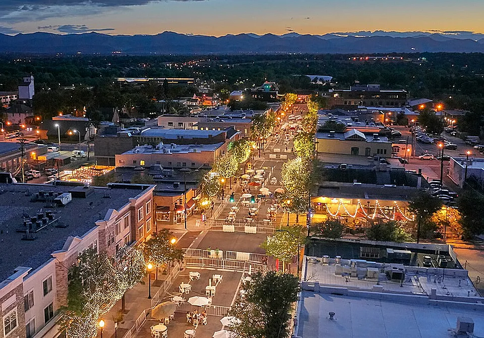 Enchanting evening aerial view of a lively city street lined with twinkling lights, bustling cafes, and picturesque mountain backdrop.