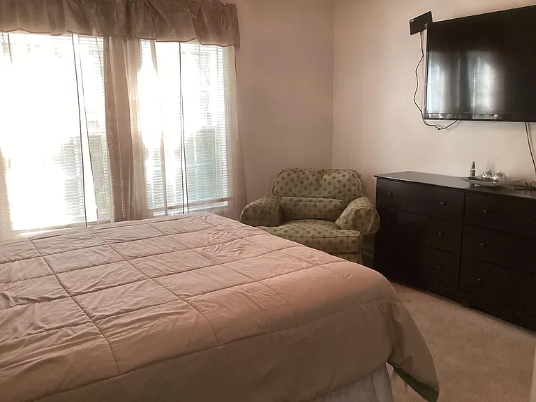 Bedroom features a large window with blinds, a comfortable bed, dresser storage, and a TV for easy in-room relaxing.