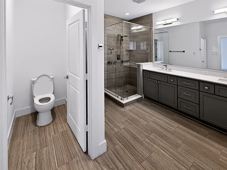 Spacious bathroom featuring wood-finish tile flooring, a dual vanity with dark cabinetry and light countertops, a large frameless mirror, and a glass-enclosed shower with tiled walls