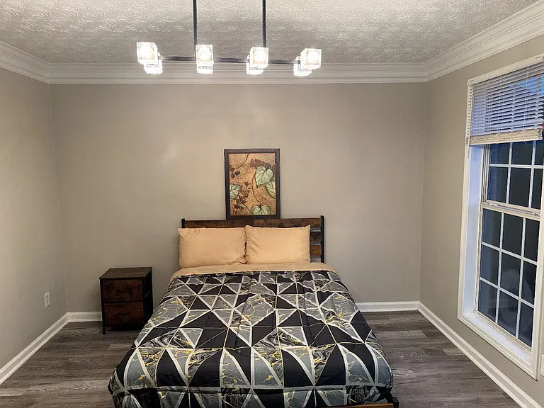 Private bedroom with a full bed, nightstand, and large window with blinds, plus durable flooring and overhead lighting.
