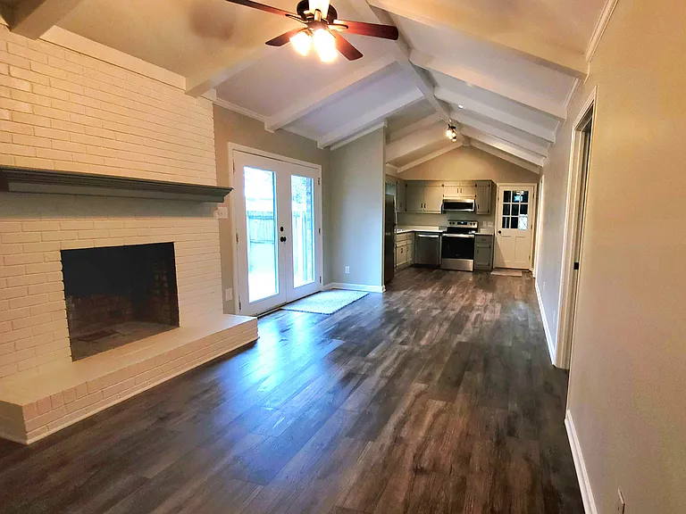 View of the den and the kitchen. Fireplace is decorative only. Room features wood/laminate flooring, french doors to the outside patio/backyard, beamed ceilings, neutral colors.