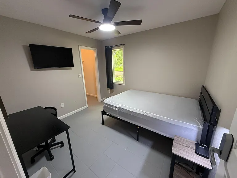 Furnished bedroom featuring a ceiling fan, window with curtains, wall-mounted TV, and a dedicated desk workspace.