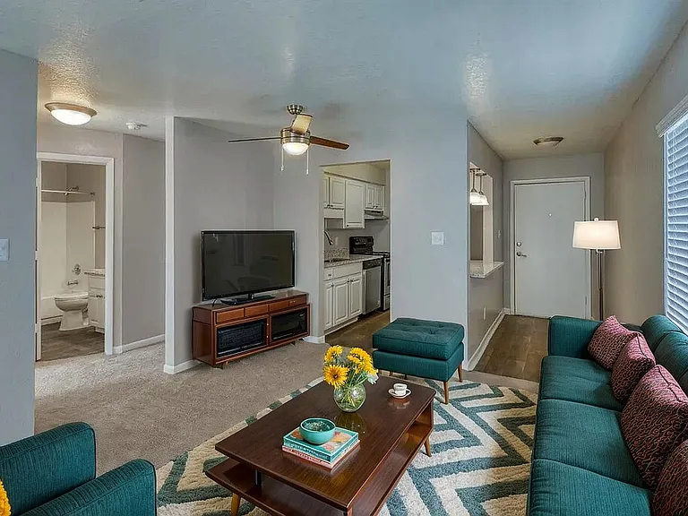 Inviting living room with modern teal sofa, bright accent pillows, and stylish coffee table with fresh flowers. Open layout leading to a fully-equipped kitchen and bathroom, highlighting comfort and elegance.