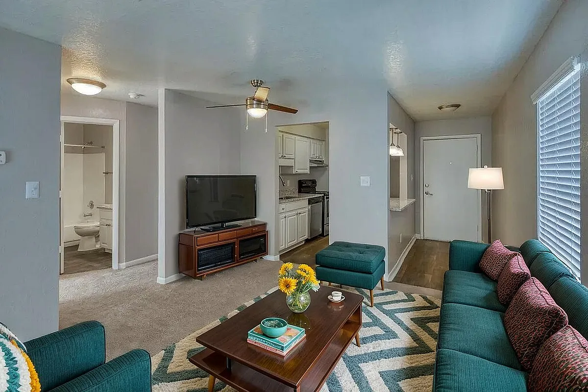 Inviting living room with modern teal sofa, bright accent pillows, and stylish coffee table with fresh flowers. Open layout leading to a fully-equipped kitchen and bathroom, highlighting comfort and elegance. - Heritage Cove