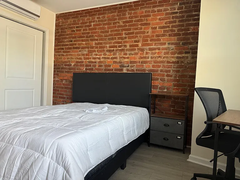 Private bedroom with exposed brick accent wall, bed, nightstand, and a dedicated desk area for work or study.