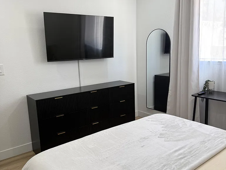 Furnished bedroom with wall-mounted TV, dresser storage, full-length mirror, and window with curtains.