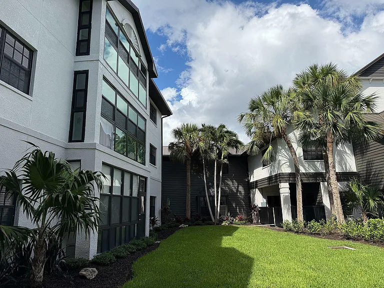 Charming apartment exterior with lush green lawn, elegant windows, and tropical palm trees under a vibrant blue sky.