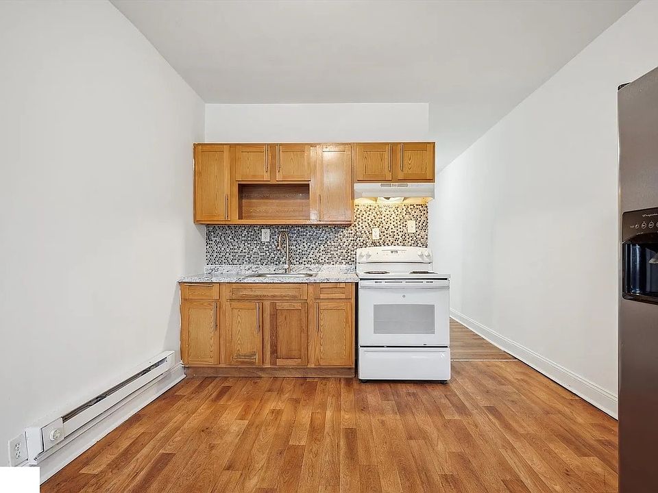Open kitchen and dining area with hardwood flooring and plenty of natural light. The kitchen features wooden cabinetry, a white stove, modern backsplash, and a full-size stainless steel refrigerator. The adjacent space offers room for a dining setup or fl