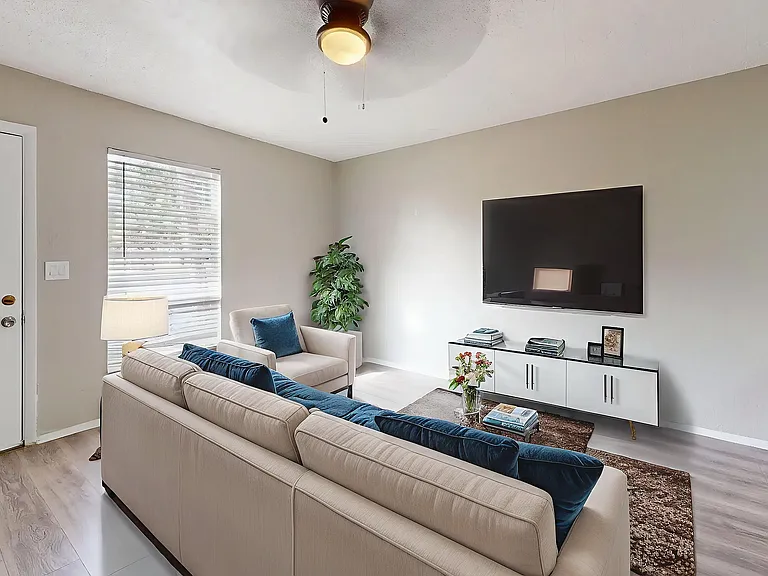 Modern living room with elegant beige sofas, a large flat-screen TV, and stylish decor. Natural light filters through a window, complementing the contemporary aesthetic and green plant accents.