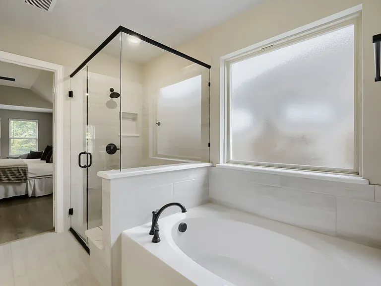 Soaking tub with matte black fixtures, glass-enclosed shower with black trim, frosted window, white subway tile surround, and light-toned wood-finish flooring