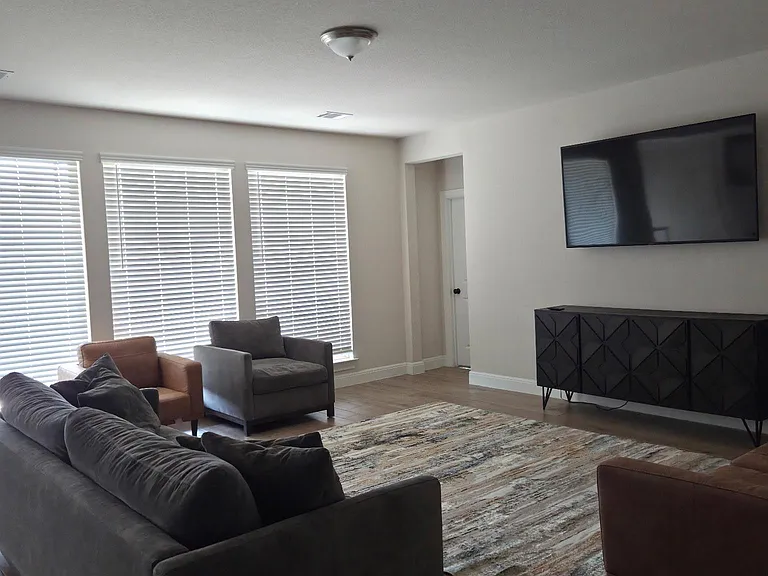 Open living room with large windows, comfortable seating, and a mounted TVideal for relaxing or gathering with housemates.
