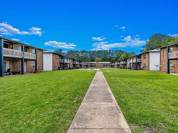 Charming apartment complex with spacious greenery and welcoming balconies, surrounded by serene trees and a bright blue sky.