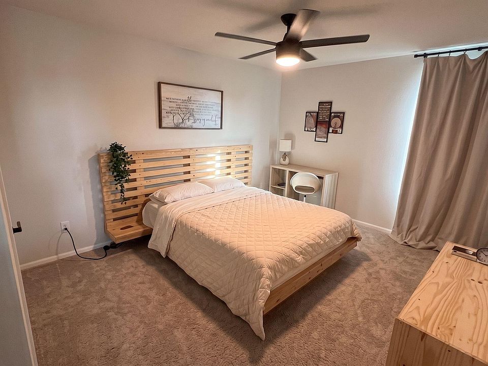 Spacious bedroom with a platform bed, ceiling fan, and soft carpeting, plus a desk nook and large window with curtains.