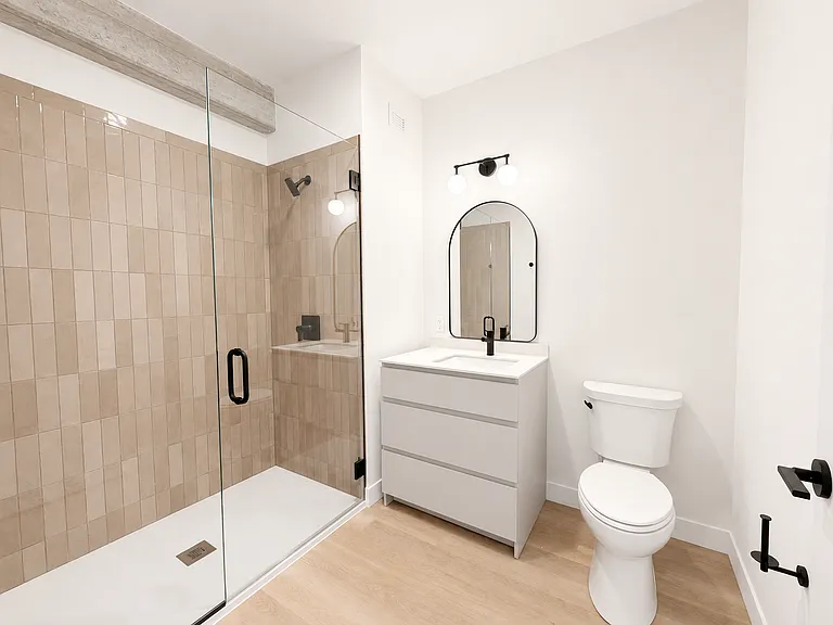 Studio bathroom at Stead 220 in Fulton Market, featuring a glass-enclosed walk-in shower with vertical subway tile, a modern white vanity, arched mirror, and matte black fixtures.