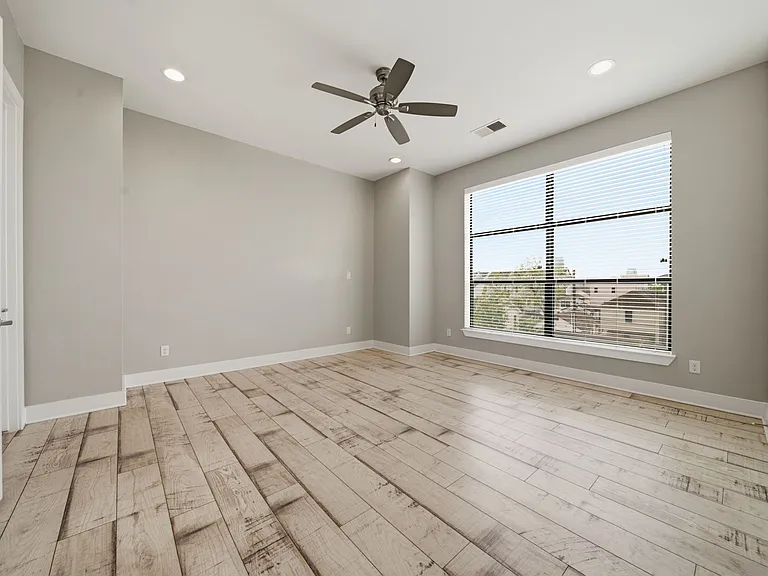 Bright and spacious primary bedroom featuring light wood flooring, neutral tones, and a large window that fills the space with natural light. Enhanced by modern ceiling fan and recessed lighting, the room also offers the convenience of dual walk-in closet